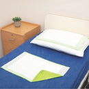 A bed with blue bedding shows two white absorbent pads with light green borders, one laid flat and one folded corner-up, alongside a white pillow. A light wood bedside table with a small potted succulent is visible in the corner.